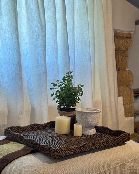 A dark brown woven square tray styled on a beige sofa, holding a small green potted plant, a white textured planter, and two cream pillar candles, set against a background of white curtains and a rustic stone column.