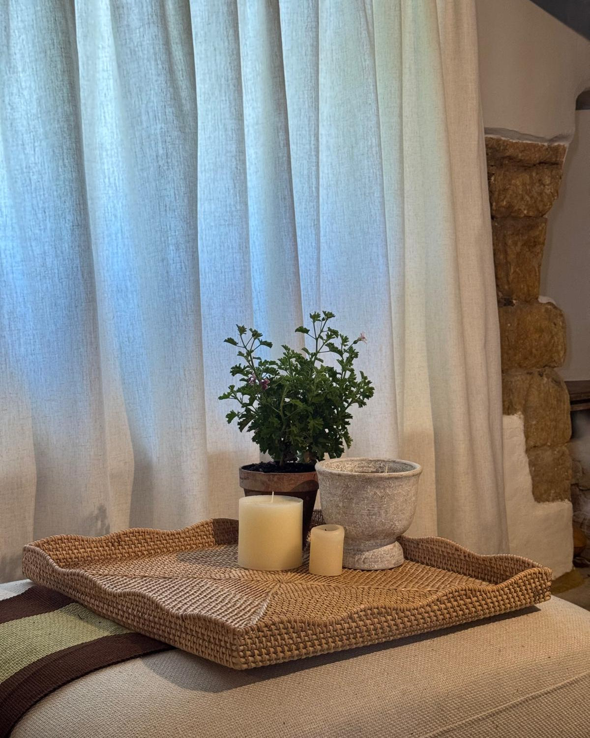A woven square tray holding a small potted green plant, a textured stone planter, and two cream candles, styled on a beige upholstered surface in front of sheer white curtains.