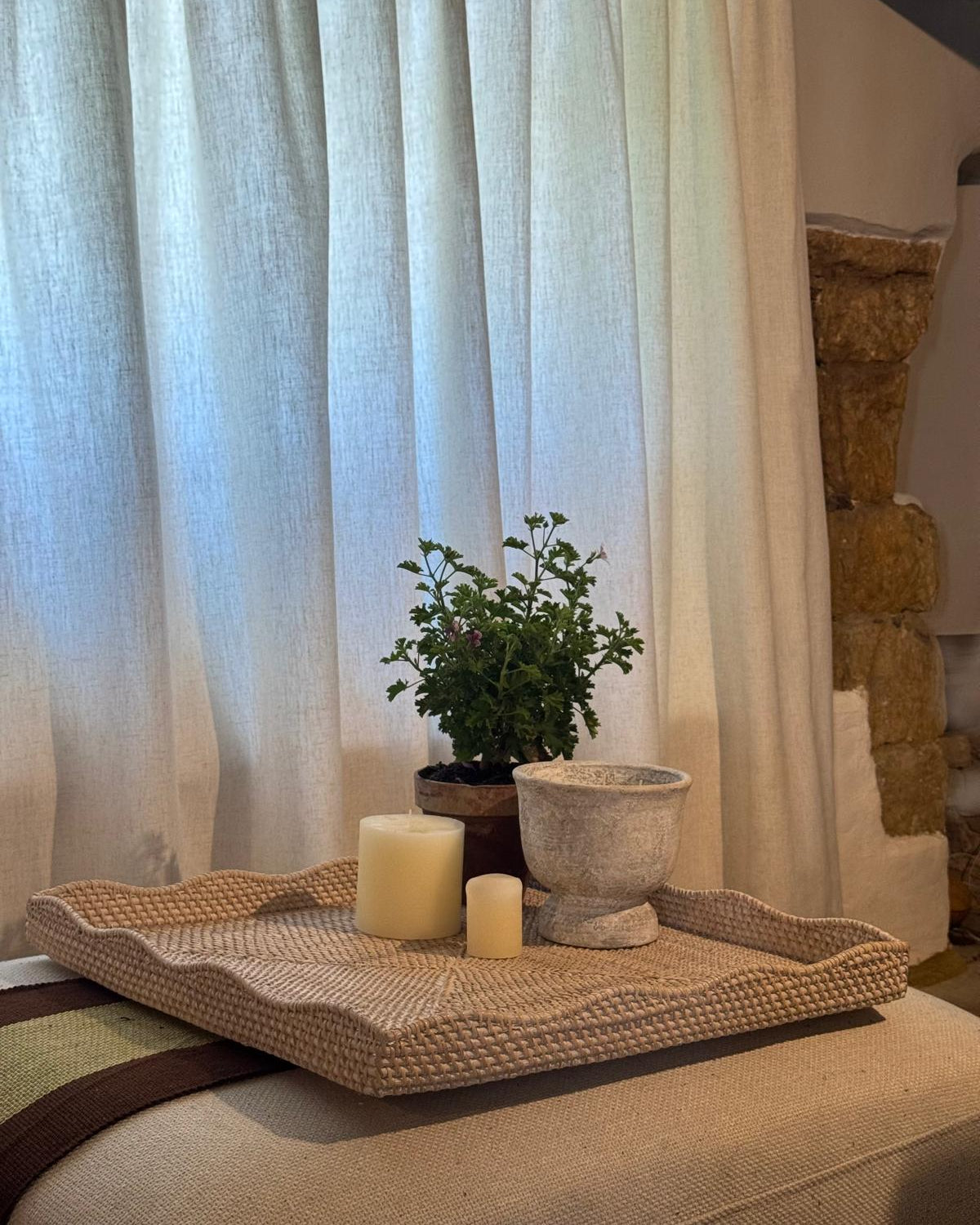 A woven natural-fiber serving tray with scalloped edges sits on a beige upholstered surface, styled with two cream candles, a small potted green herb plant, and a stone planter in front of sheer white curtains.
